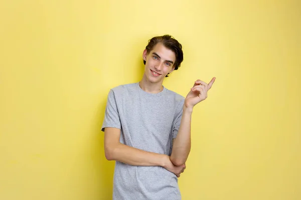 Веселый красивый молодой худой темноволосый парень с голубыми глазами в серой футболке, указывающей влево, стоя на желтом фоне — стоковое фото