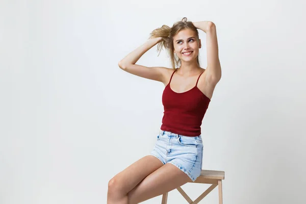 Allegro bella ragazza bionda che indossa claret t-shirt e pantaloncini di jeans blu, sorridente ridendo guardando la fotocamera, seduto su una sedia di legno in studio — Foto Stock