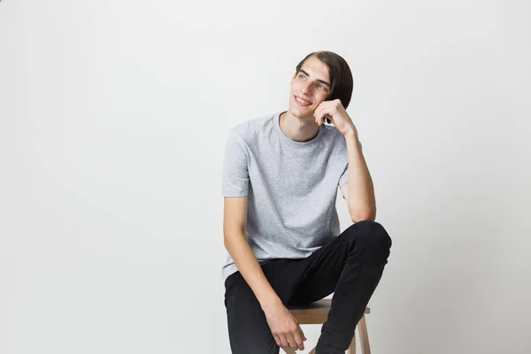 Rêveur beau jeune homme mince et brun aux yeux bleus portant un t-shirt gris, regardant vers la gauche et vers le haut, assis sur une chaise sur fond blanc — Photo