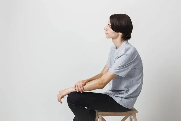 Calm and relaxed young thin dark-haired guy with blue eyes wearing gray t-shirt, sitting on chair against white background. Side view — Stock Photo, Image