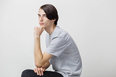 Sakin ve rahat genç ince siyah saçlı bir adam beyaz arka plan karşı sandalyeye oturan gri t-shirt giyen mavi gözlü. Yan görünüm