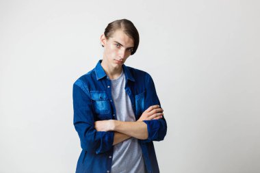 Sakin genç ince esmer yakışıklı olan mavi kot gömlek ayakta çapraz elle beyaz arka plan üzerinde mavi gözlü.