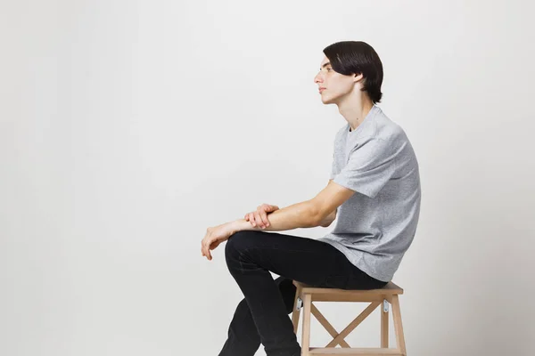 Ήρεμη και χαλαρή λεπτός μελαχρινός νεαρός με μπλε μάτια, φορώντας γκρι t-shirt, κάθεται σε καρέκλα ενάντια σε λευκό φόντο. Πλάγια όψη — Φωτογραφία Αρχείου