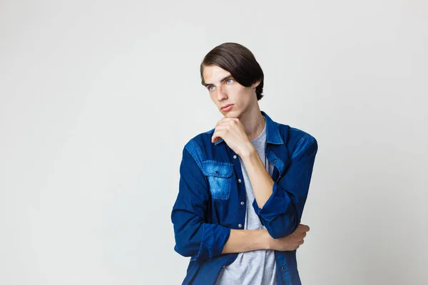 Thoughtful handsome young thin dark-haired guy with blue eyes wearing blue denim shirt, holding hand on chin, standing against white background — Stock Photo, Image