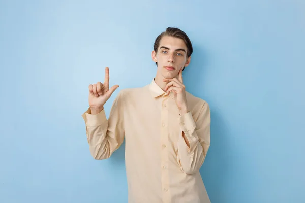 Handsome young thin dark-haired guy with blue eyes wearing yellow shirt pointing up while posing over blue wall