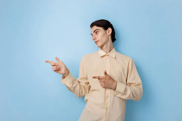 Guapo joven delgado de pelo oscuro chico con ojos azules usando camisa amarilla apuntando a la izquierda mientras posando sobre la pared azul — Foto de Stock