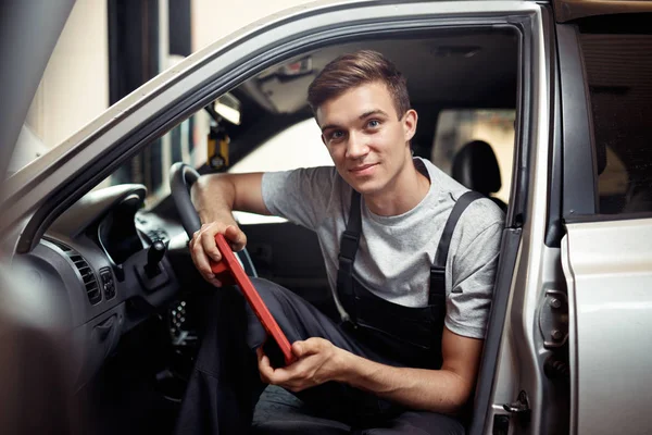 Un jeune mécanicien souriant est assis dans une voiture avec un dispositif spécial pour cheching le système informatique d'une voiture — Photo