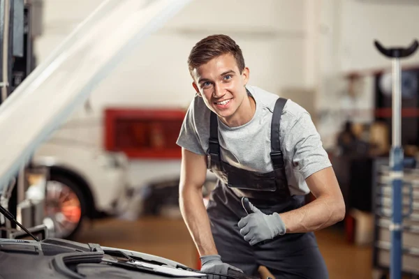 Un joven sonríe de pie cerca de un coche en su trabajo. Mantenimiento de automóviles y vehículos — Foto de Stock