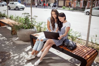 Güneşli bir öğleden sonra uzun siyah saçlı, iki genç güzel bayanlar giyiyor rahat slothes, birbirine yakın bankta oturmak ve dikkatle bakmak belgili tanımlık screenâ.
