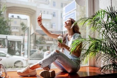 Uzun saç, rahat kıyafetler giymiş güzel bir genç kızla bir pencere üzerinde oturur ve bir selfie bir fincan kahve ile modern bir kafede alır.