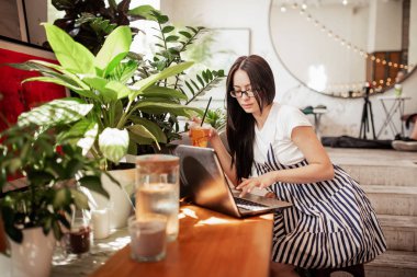 Güzel bir genç ince kız siyah saçlı ve gözlük, casual tarzda giyinmiş ile bir dizüstü bilgisayar yeni bir kafede masada oturur.