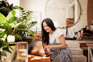 Güzel bir genç ince kız siyah saçlı ve gözlük, casual tarzda giyinmiş ile bir dizüstü bilgisayar yeni bir kafede masada oturur.