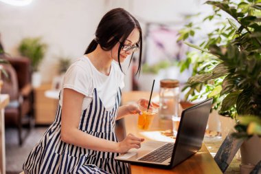 Güzel bir genç ince kız siyah saçlı ve gözlük, casual tarzda giyinmiş ile bir dizüstü bilgisayar yeni bir kafede masada oturur.