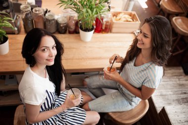Koyu saçlı, gündelik kıyafeti giymiş iki güzel genç gülümseyen kız karşılıklı oturup rahat bir kafede sohbet.