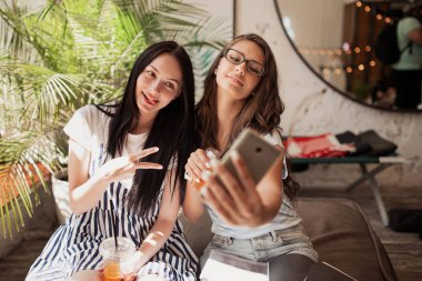 İki genç gülümseyen güzel rahat kıyafetler giyen kızlar uzun siyah saçlı, ince yapılı, yan yana oturup bir selfie modern bir kahve dükkanında.