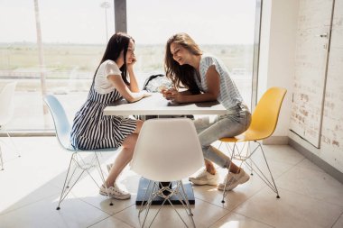 Koyu saçlı, iki genç oldukça ince kız gündelik kıyafeti giymiş birbirine yanındaki masada ve modern bir ofiste sohbet.
