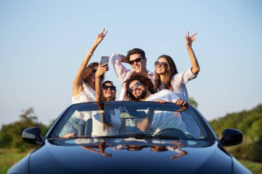 Neşeli genç kızlar ve çocuklar güneş gözlüğü ellerini tutarak ve selfie güneşli bir günde yapma yolu üzerinde siyah bir Cabriolet oturuyor.
