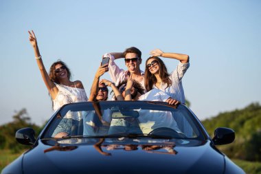 Komik genç kızlar ve çocuklar güneş gözlüğü ellerini tutarak ve selfie güneşli bir günde yapma yolu üzerinde siyah bir Cabriolet oturuyor.
