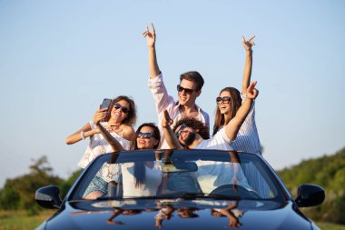 Güzel genç kızlar ve çocuklar güneş gözlüğü ellerini tutarak ve selfie güneşli bir günde yapma yolu üzerinde siyah bir Cabriolet oturuyor.