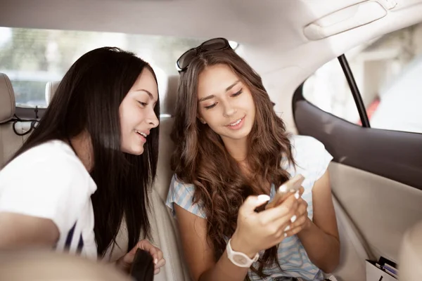 Two youthful friendly dark-haired girls,dressed in casual style, are sitting in the backseat of a fancy car and showing something to each other. — Stock Photo, Image