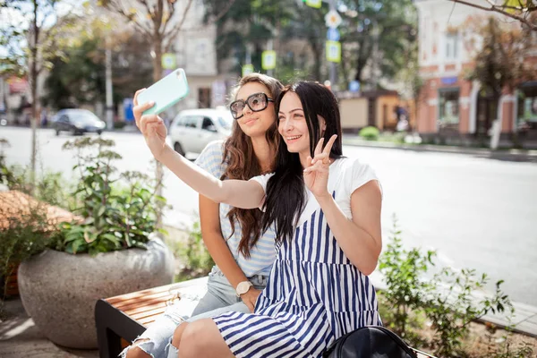 Két jó látszó vékony lány hosszú, Sötét haja, alkalmi disznóól, öltözött ül a padon, és vesz egy selfie, — Stock Fotó