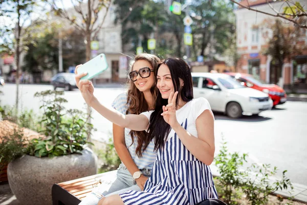 Due belle ragazze magre con lunghi capelli scuri, vestite in stye casual, sedersi in panchina e farsi un selfie , — Foto Stock