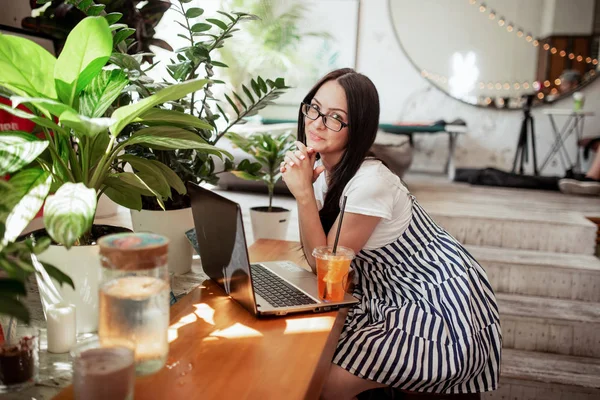Красива молода струнка дівчина з темним волоссям і окулярами, одягнена в казуальний стиль, сидить за столом з ноутбуком і дивиться на камеру в затишній кав'ярні . — стокове фото