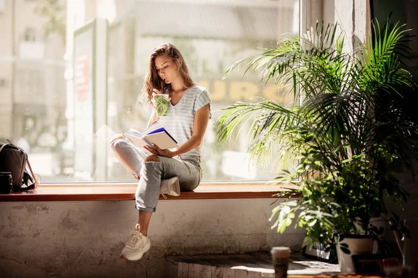 Une jolie jeune fille mince aux cheveux longs, vêtue d'une tenue décontractée, assise sur le rebord de la fenêtre et boit du café et lit un livre dans un café confortable . — Photo