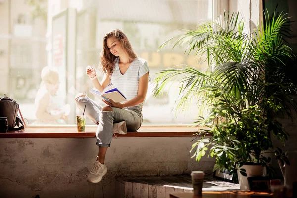 Uma jovem muito magra com cabelos longos, vestindo roupas casuais, sentar-se no peitoril da janela e lê um livro em um café acolhedor . — Fotografia de Stock