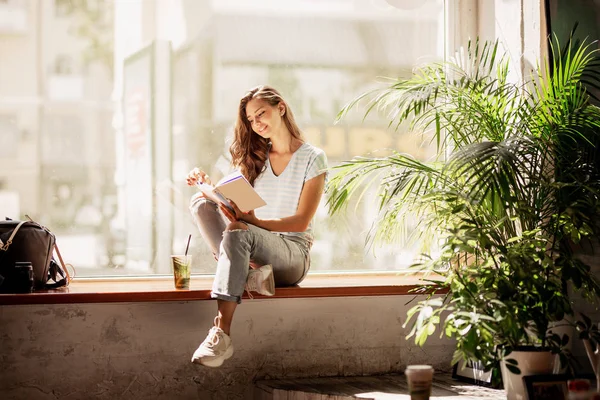 Ein hübsches, schlankes, junges Mädchen mit langen Haaren, lässig gekleidet, sitzt auf der Fensterbank und liest in einem gemütlichen Café ein Buch. — Stockfoto