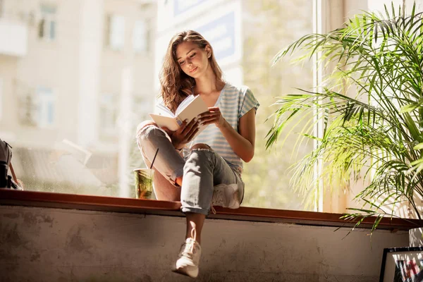 Довольно стройная девушка с длинными волосами, в повседневной одежде, сидит на подоконнике и читает книгу в уютном кафе . — стоковое фото