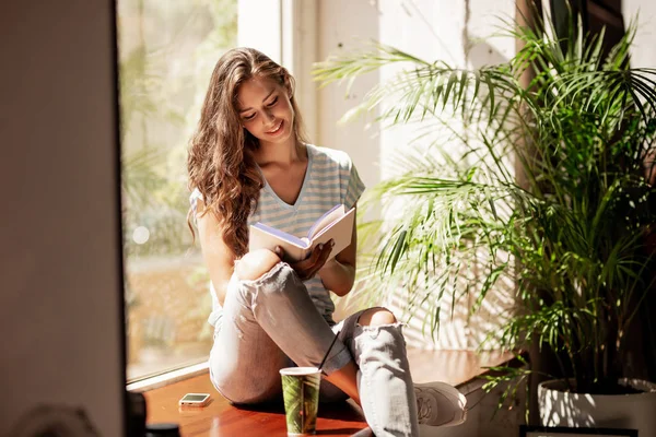 Une jolie jeune fille mince aux cheveux longs, portant une tenue décontractée, assise sur le rebord de la fenêtre et lit un livre dans un café confortable . — Photo