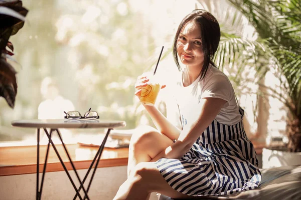 Uma jovem menina sorridente magro com cabelo escuro, vestido com roupa casual, senta-se à mesa e bebe café em uma loja de café acolhedora . — Fotografia de Stock