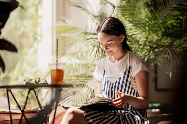 Una giovane ragazza magra e amichevole con i capelli scuri, vestita con abiti casual, si siede a tavola e legge un libro in un'accogliente caffetteria. . — Foto Stock