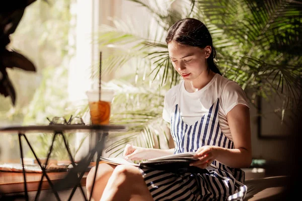Молода струнка дружня дівчина з темним волоссям, одягнена в повсякденне вбрання, сидить за столом і читає книгу в затишній кав'ярні . — стокове фото