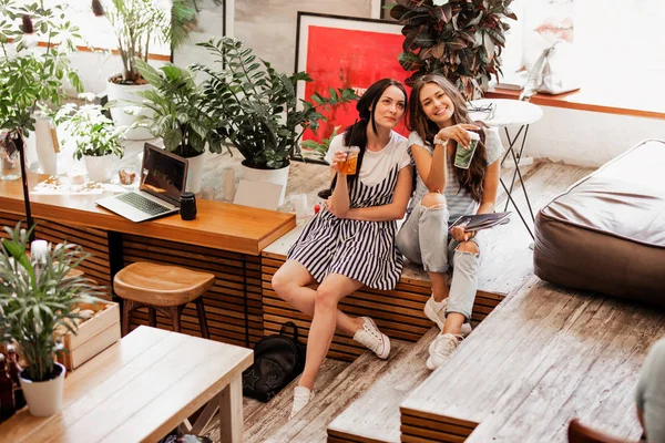 Twee jonge lachende meisjes met lang donker haar, het dragen van casual outfit, zitten naast elkaar en drinken koffie in een gezellige coffee shop. — Stockfoto