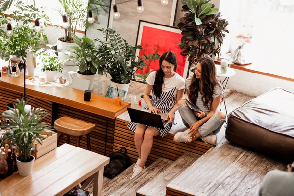 เด็กสาวสองคนที่ยิ้ม มีผมดํายาว ใส่ชุดลําลอง นั่งอยู่ข้างๆกัน และดื่มกาแฟในร้านกาแฟที่อบอุ่น . — ภาพถ่ายสต็อก