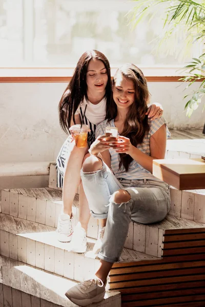Дві молоді красиві дівчата з довгим темним волоссям, одягнені в повсякденне вбрання, сидять на сходах і дивляться на телефон у затишній кав'ярні . — стокове фото