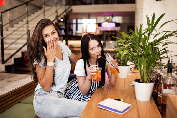 Twee vrij jeugdige lachende meisjes, gekleed in een casual outfit, naast elkaar zitten en kijken naar de camera in een gezellige coffee shop. — Stockfoto