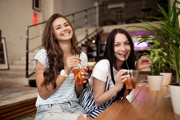 Dos chicas sonrientes bastante jóvenes, vestidas con ropa casual, se sientan una al lado de la otra y miran a la cámara en una acogedora cafetería . —  Fotos de Stock