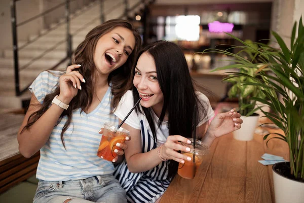 Δύο αρκετά νεανική χαμογελαστών κοριτσιών, ντυμένοι με casual ντύσιμο, κάθονται δίπλα στο άλλο και να εξετάσουμε την κάμερα σε ένα άνετο κατάστημα καφέ. — Φωτογραφία Αρχείου