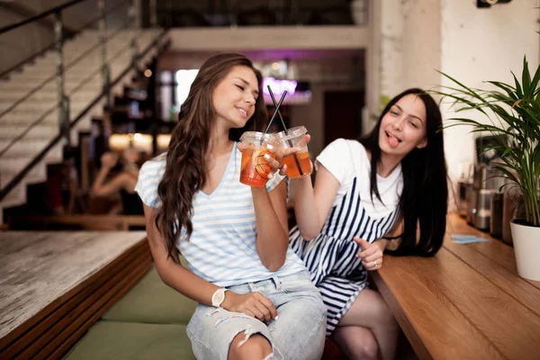 Дві красиві молоді усміхнені дівчата, одягнені в повсякденне вбрання, сидять поруч один з одним і дивляться на камеру в затишній кав'ярні . — стокове фото