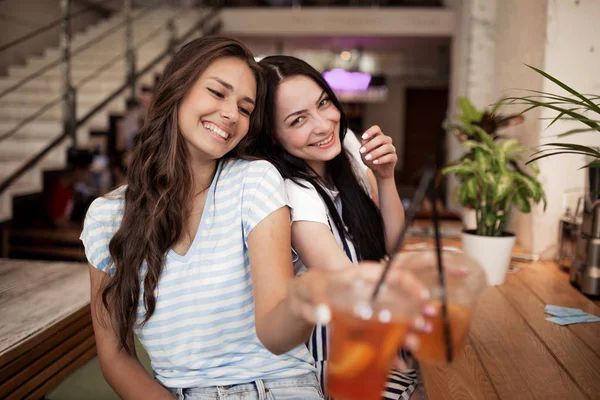 Deux jolies jeunes filles souriantes, vêtues d'une tenue décontractée, s'assoient l'une à côté de l'autre et regardent la caméra dans un café confortable . — Photo