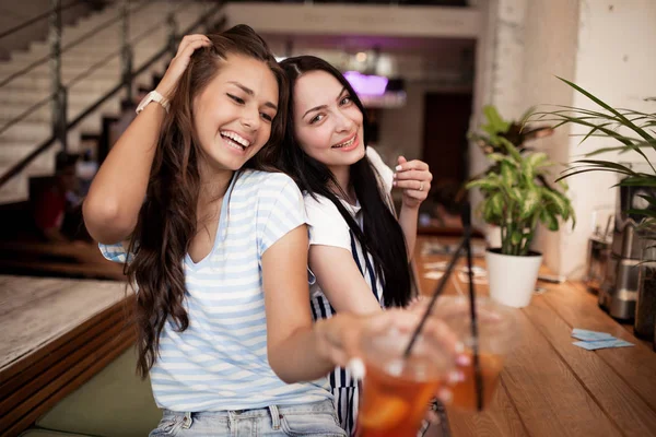 Twee vrij jeugdige lachende meisjes, gekleed in een casual outfit, naast elkaar zitten en kijken naar de camera in een gezellige coffee shop. — Stockfoto