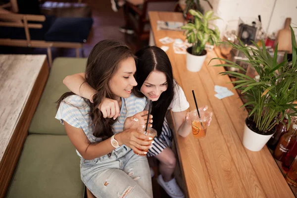 Zwei schöne junge lächelnde Mädchen mit dunklen Haaren, in lässigem Outfit gekleidet, umarmen einander in einem gemütlichen Café. — Stockfoto