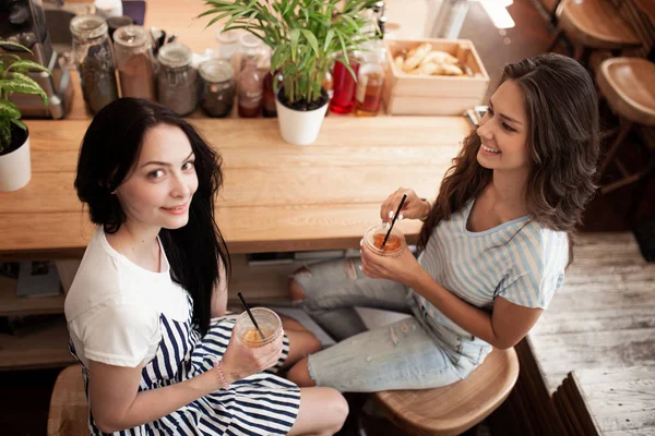 Duas belas garotas sorridentes jovens com cabelo escuro, vestidas com roupas casuais, sentam-se em frente uma da outra e conversam em um aconchegante café. . — Fotografia de Stock