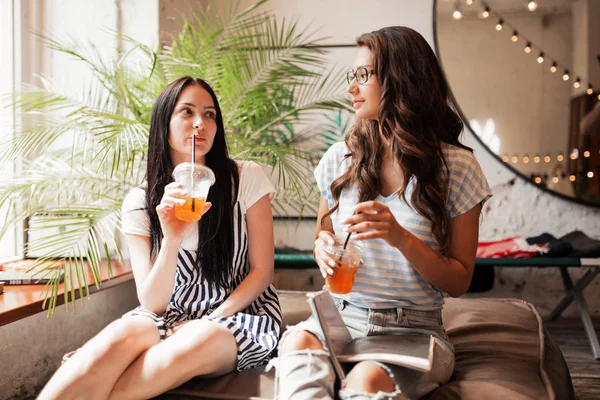 Дві молоді красиві дівчата з довгим темним волоссям, одягнені в повсякденний одяг, сидять поруч один з одним і п'ють каву в сучасному кафе . — стокове фото