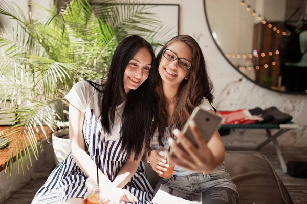 Дві молоді усміхнені красиві стрункі дівчата з довгим темним волоссям, одягнені в повсякденний одяг, сидять поруч один з одним і приймають селфі в сучасному кафе . — стокове фото