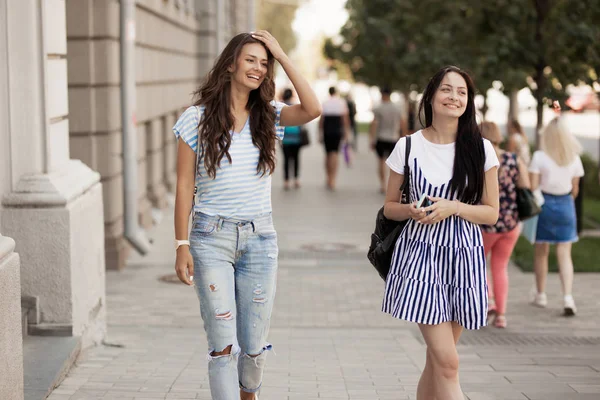 Deux jeunes filles minces et mignonnes aux cheveux longs, marchent dans la rue par une journée ensoleillée . — Photo