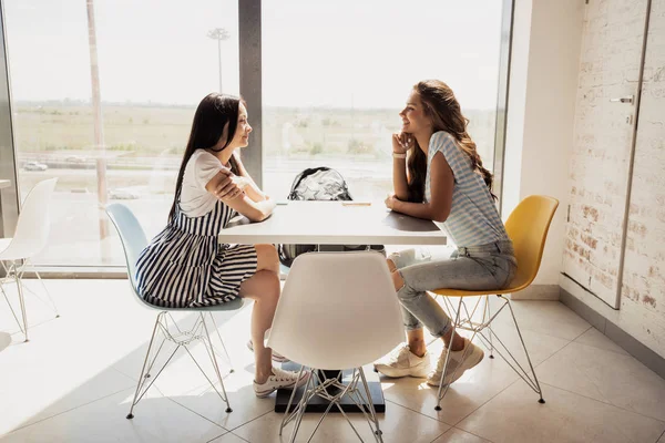 Două fete tinere destul de subțiri, cu păr întunecat, purtând costum casual, stau la masă unul lângă celălalt și vorbesc într-un birou modern . — Fotografie, imagine de stoc